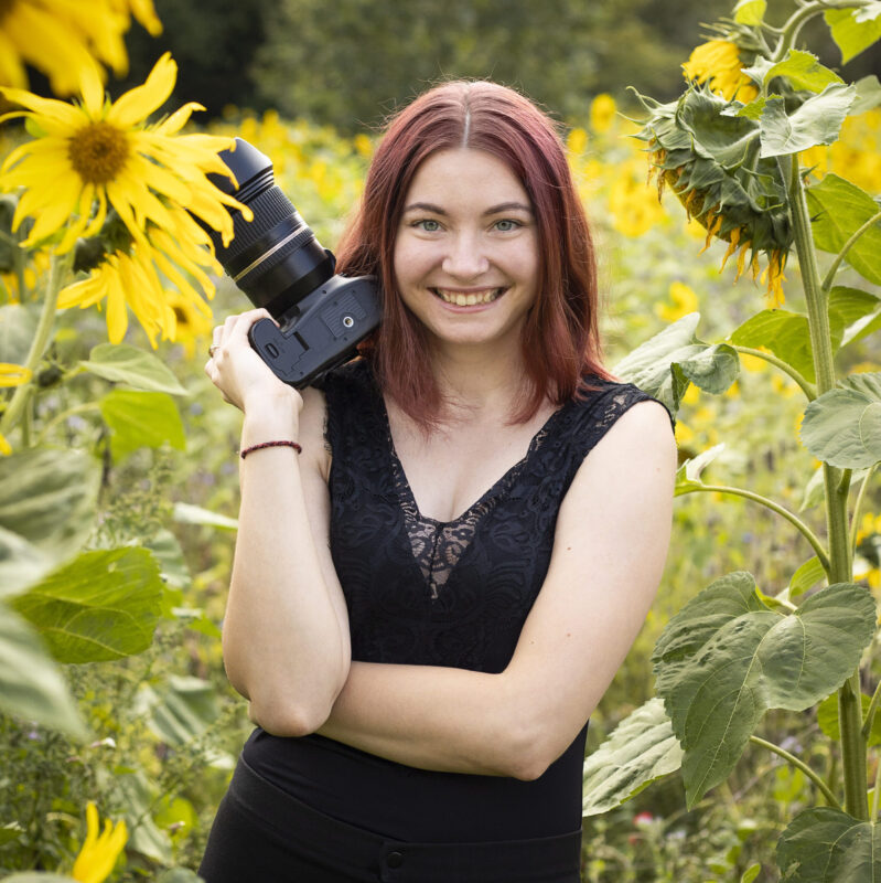 Rothaarige Frau steht mit ihrer Kamera in einem Sonnenblumenfeld und lacht