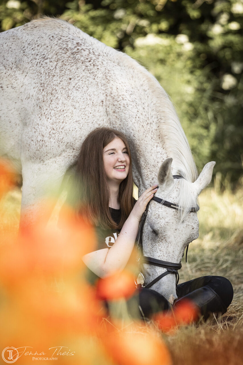 Junges Mädchen sitzt auf dem Boden und lacht, ihr weißes Pferd beugt den Kopf in ihrem Schoß, im Vordergrund sind unscharfe rote Mohnblumen