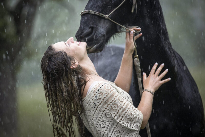 Lachende Frau legt den Kopf zurück und kuschelt mit ihrem Friesen im Regen