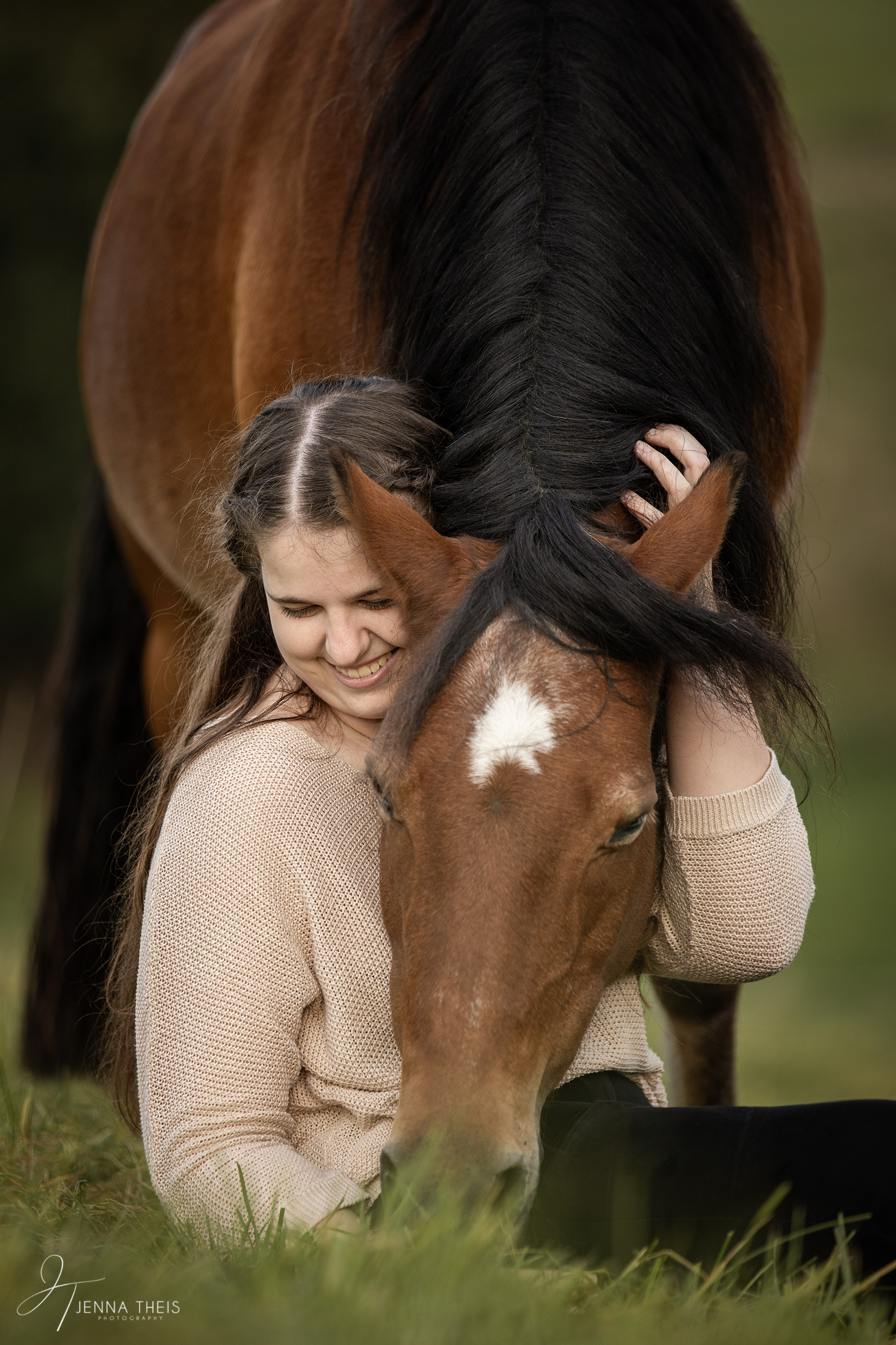 Lachendes Mädchen sitzt auf einer Wiese, Pony hat seinen Kopf in ihrem Schoß