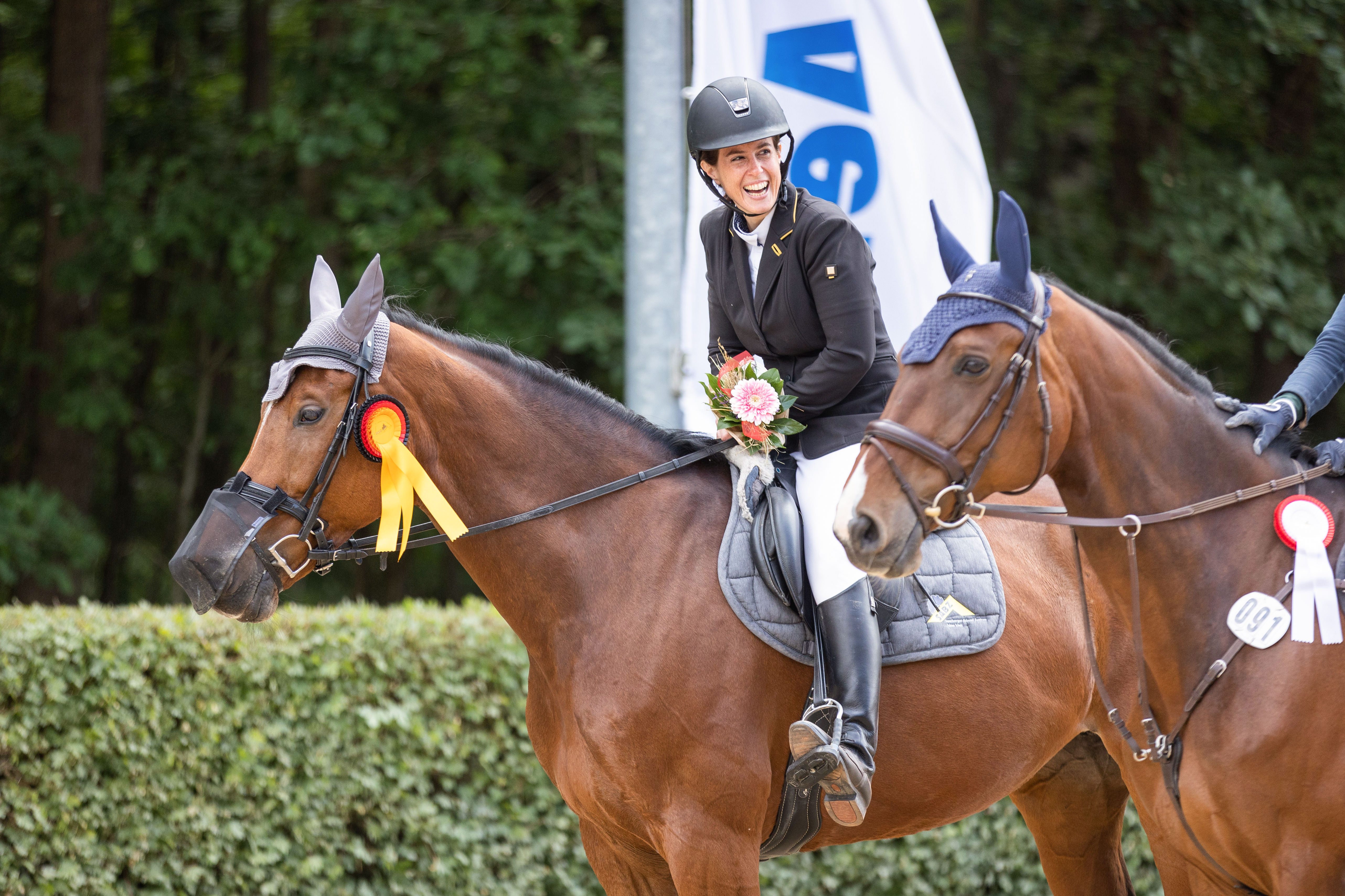 Lachende Frau mit Blumen in der Hand sitzt auf ihrem Pferd während der Siegerehrung von einem Springturnier