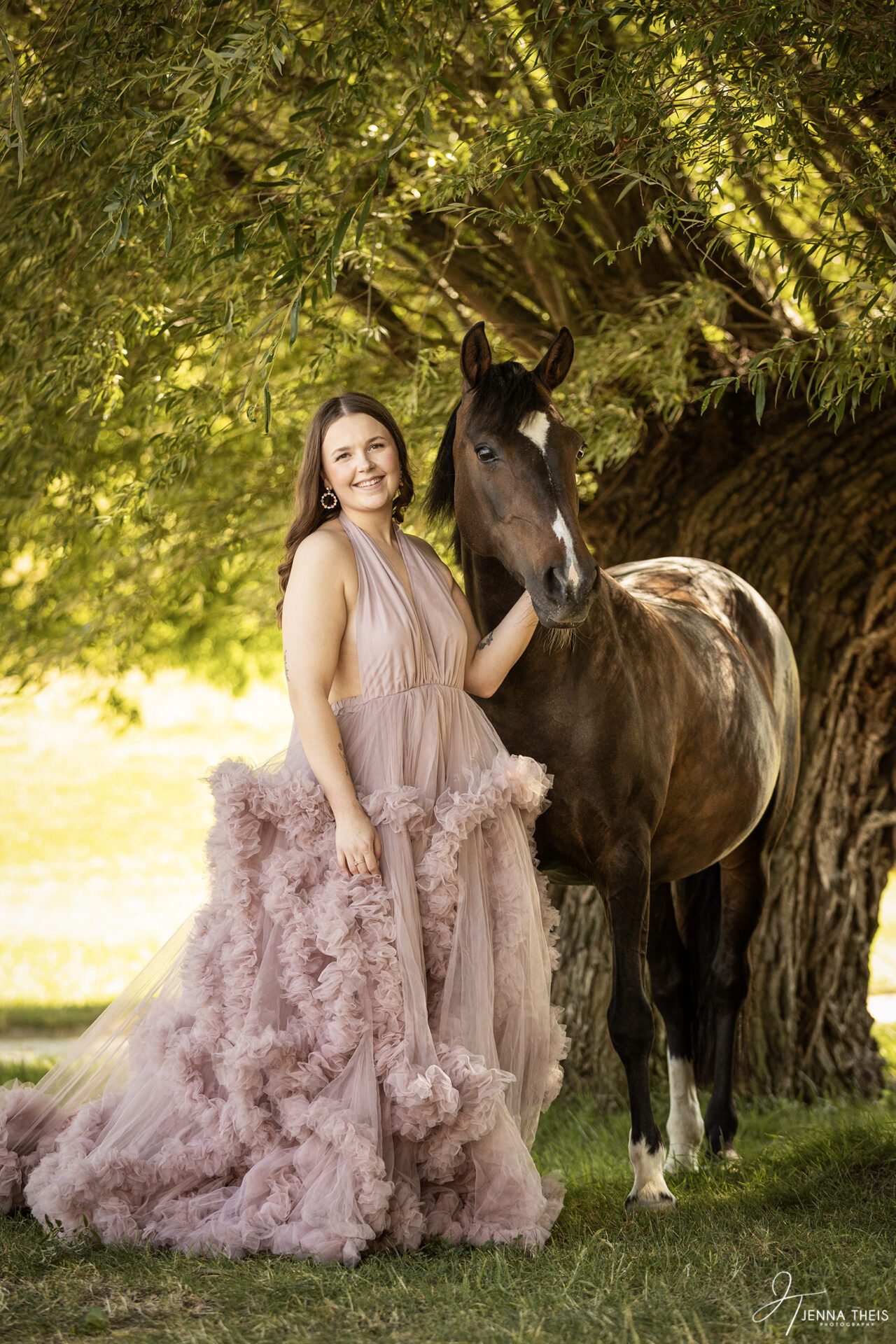 Frau in langem rosa Kleid steht neben ihrem braunen Pony vor einem Baum