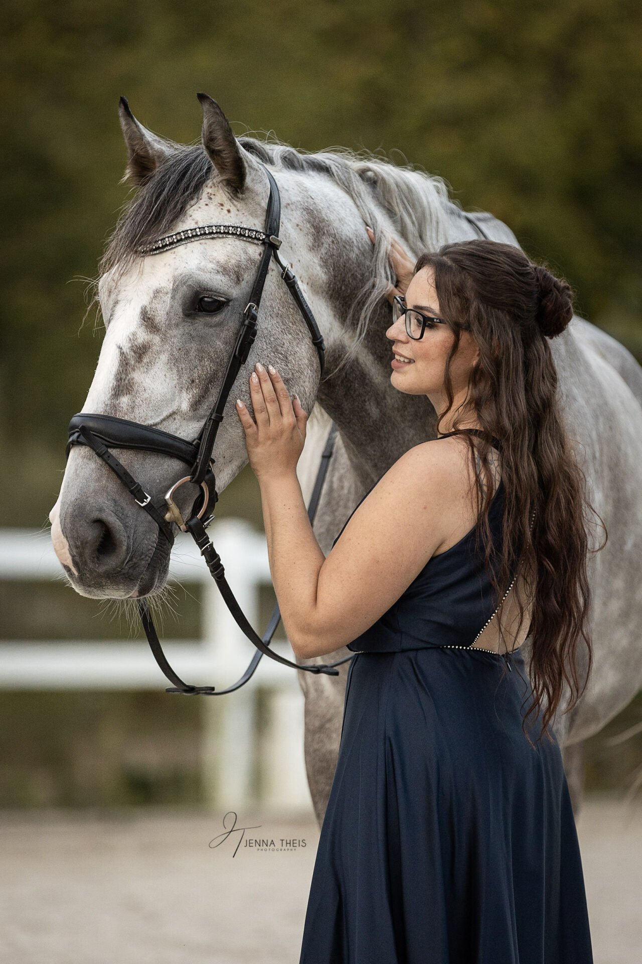 Frau in blauem Kleid steht auf einem Reitplatz neben ihrem Apfelschimmel