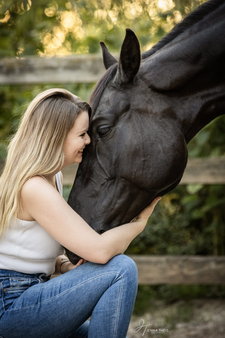 Junge Frau in der Hocke kuschelt mit ihrem schwarzen Pferd